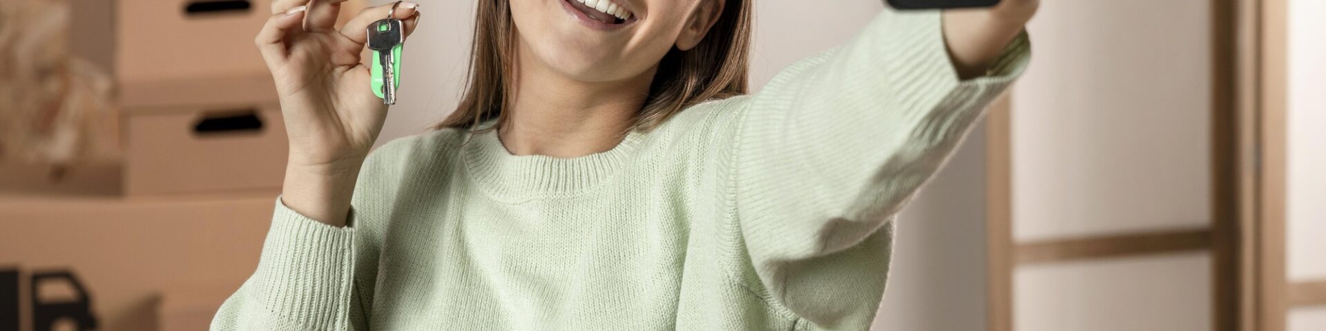 mulher tirando uma selfie segurando uma chave e sorrindo para a foto. Ao fundo, caixas de mudança.